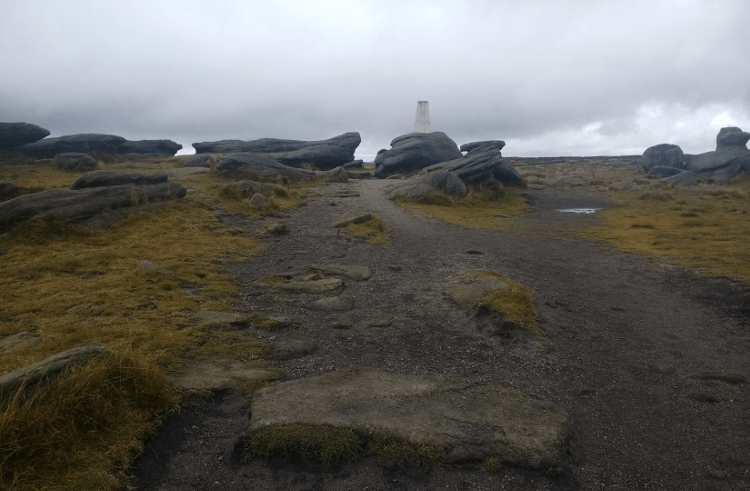 Looking up at a trig point across peaty land.